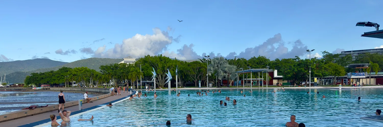 Swimmers in the Cairns Esplanade Lagoon with palm trees, parkland and green hills behind under a blue sky
